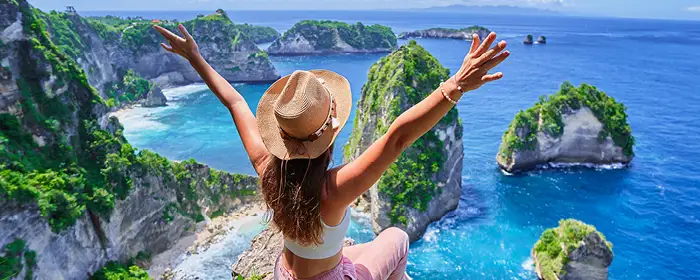 Women sitting on an cliff edge in a tropical paradise looking over the ocean with her arms in the air